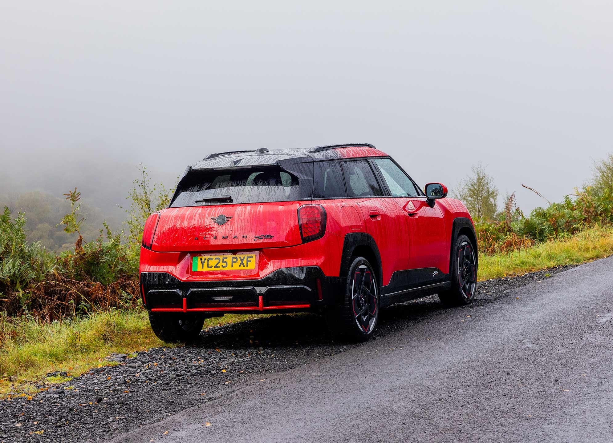 Rear side view of parked all-electric MINI Aceman in British Racing Green in car park with all doors open and coloured wall in the background